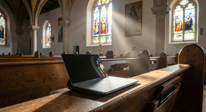 A laptop on a wooden pew showing a clean church home page in sunlight from a stained glass window.